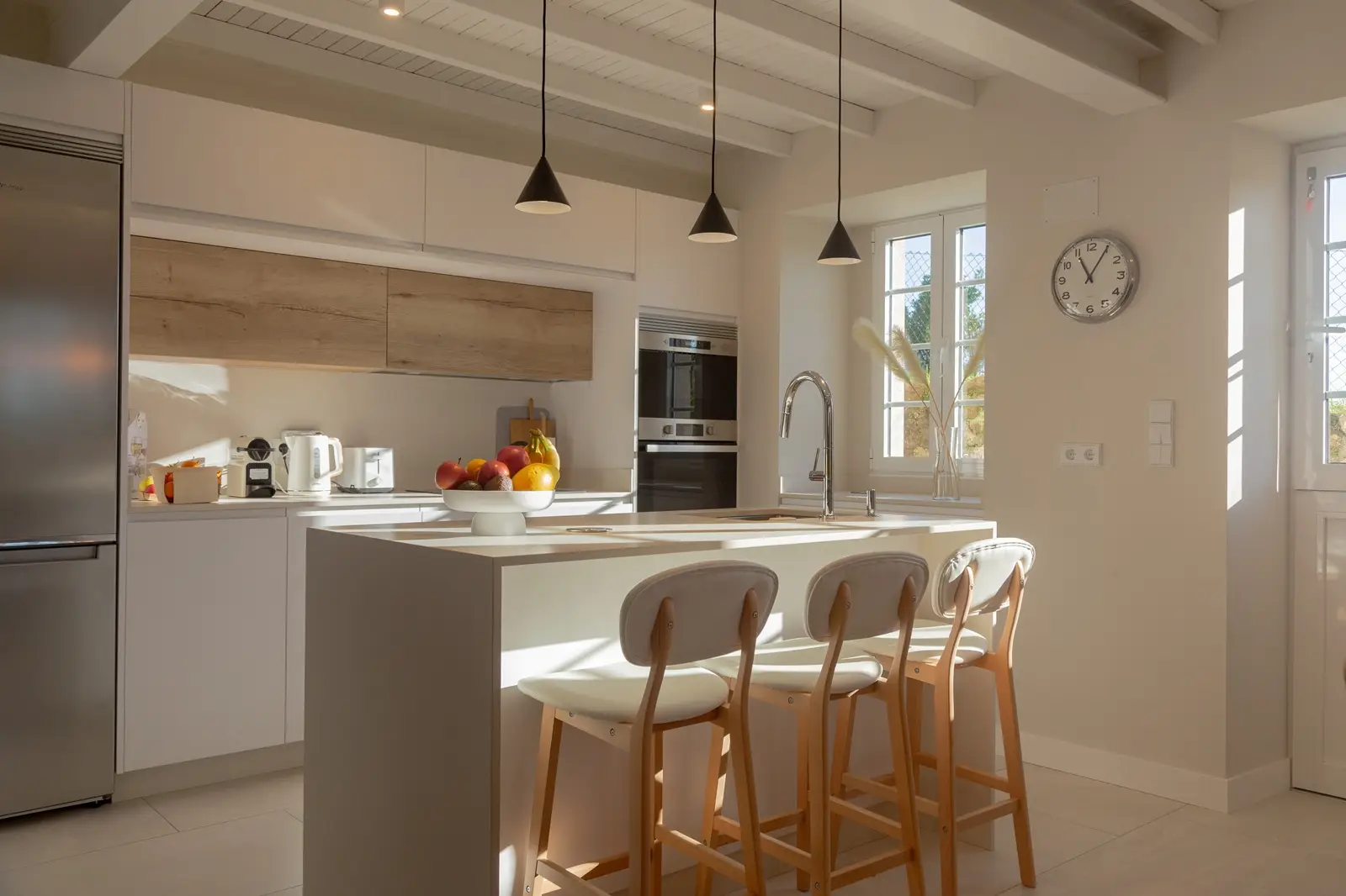 Cocina moderna con barra iluminada por luz natural que entra por la ventana, acompañada de tres sillas de barra beige
