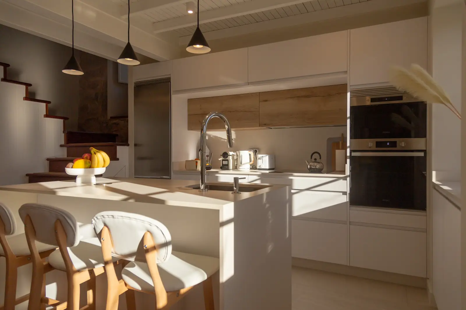 Cocina moderna con barra iluminada por luz natural que entra por la ventana, acompañada de tres sillas de barra beige