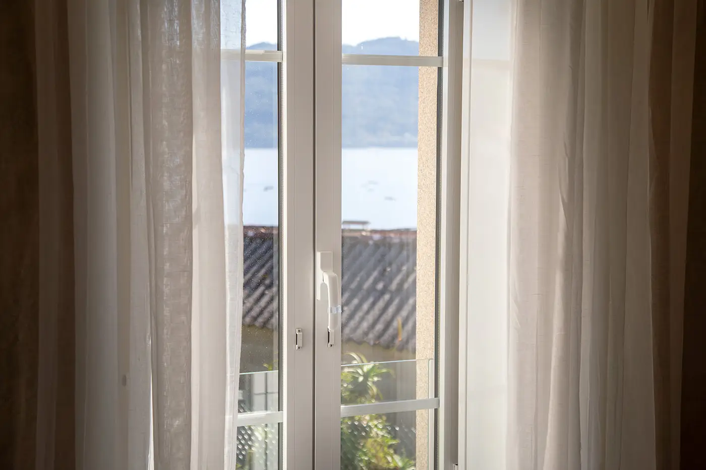 acercamiento a ventana cerrada con cortinas blancas desde la cual se ve el paisaje gallego y el mar