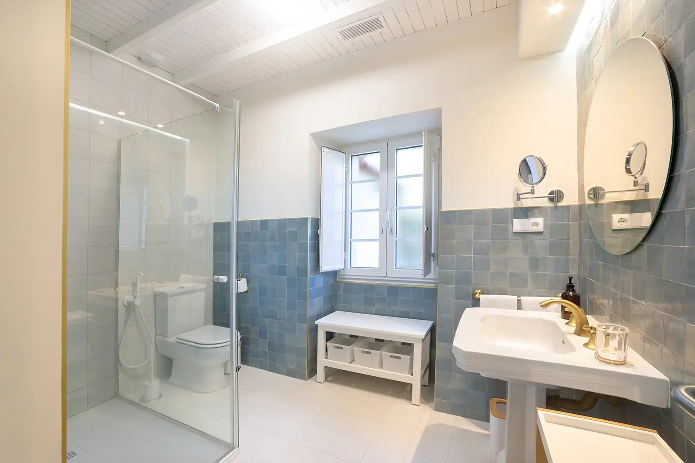 Vista general del baño con azulejos color azul zona de almacenamiento, espejo grande redondo, lavamanos y zona de ducha